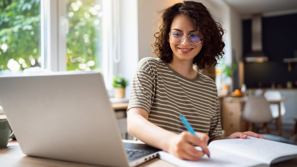 Young woman writing notes with her laptop open in front of her