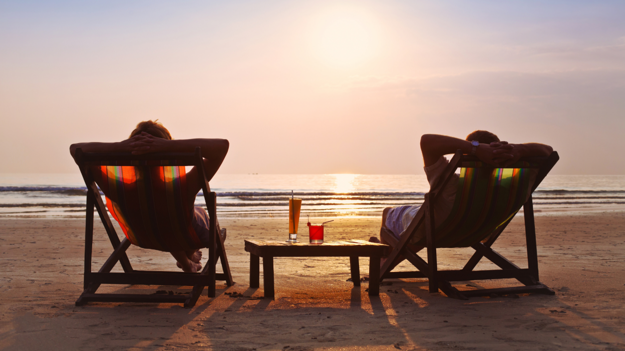 A couple relaxing in chairs on the beach with two cocktails on a table between them