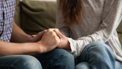 A closeup of a man and woman sitting on a couch, visible from the chest down and holding both. of one another's hands