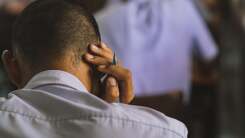 A young man sits facing away from the camera and holding his head as if stressed