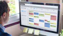 A man looking at a calendar app on his desktop computer, with his schedule filled with colored squares