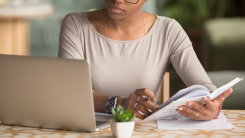 A woman at a table studying from a textbook with a laptop open in front of her