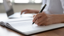 A person writing with pen and paper in front of a laptop