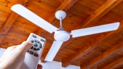 hand pointing remote control at white ceiling fan in front of wood ceiling