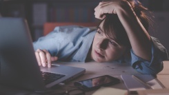 A photograph of a woman leaning onto a desk while she uses a laptop, looking sleepy