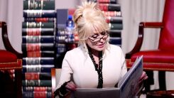 Dolly Parton reading a children's book on stage with a backdrop of books