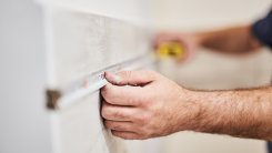 A person's hands using a metal tape measure to evaluate the length of a wall.