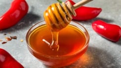 A honey dipper drizzling honey into a bowl with red peppers in the background.