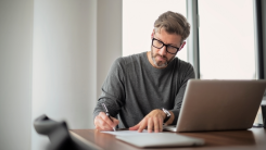 man working with a laptop and writing down notes on paper