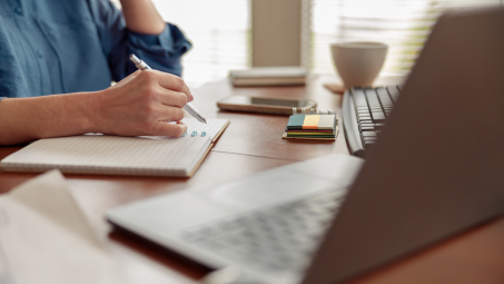 person writing in a notebook with laptop computer open to the side