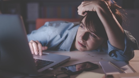 A photograph of a woman leaning onto a desk while she uses a laptop, looking sleepy